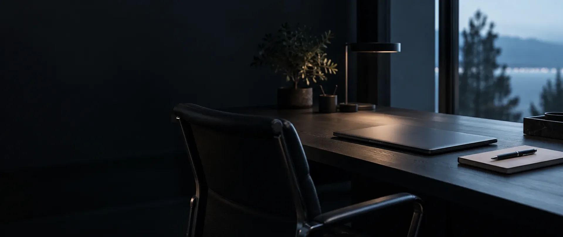 Quiet dark workspace with an empty chair, closed laptop, notebook and pen beside a window