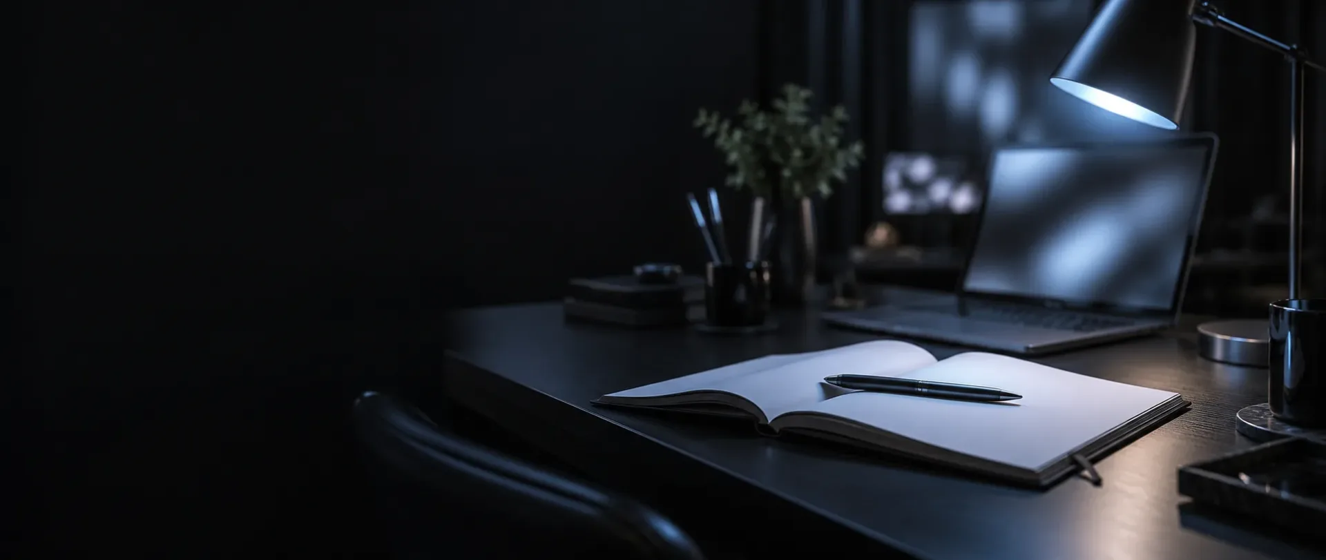 Dark writing desk with an open blank notebook and pen, a desk lamp and a laptop in soft blue light
