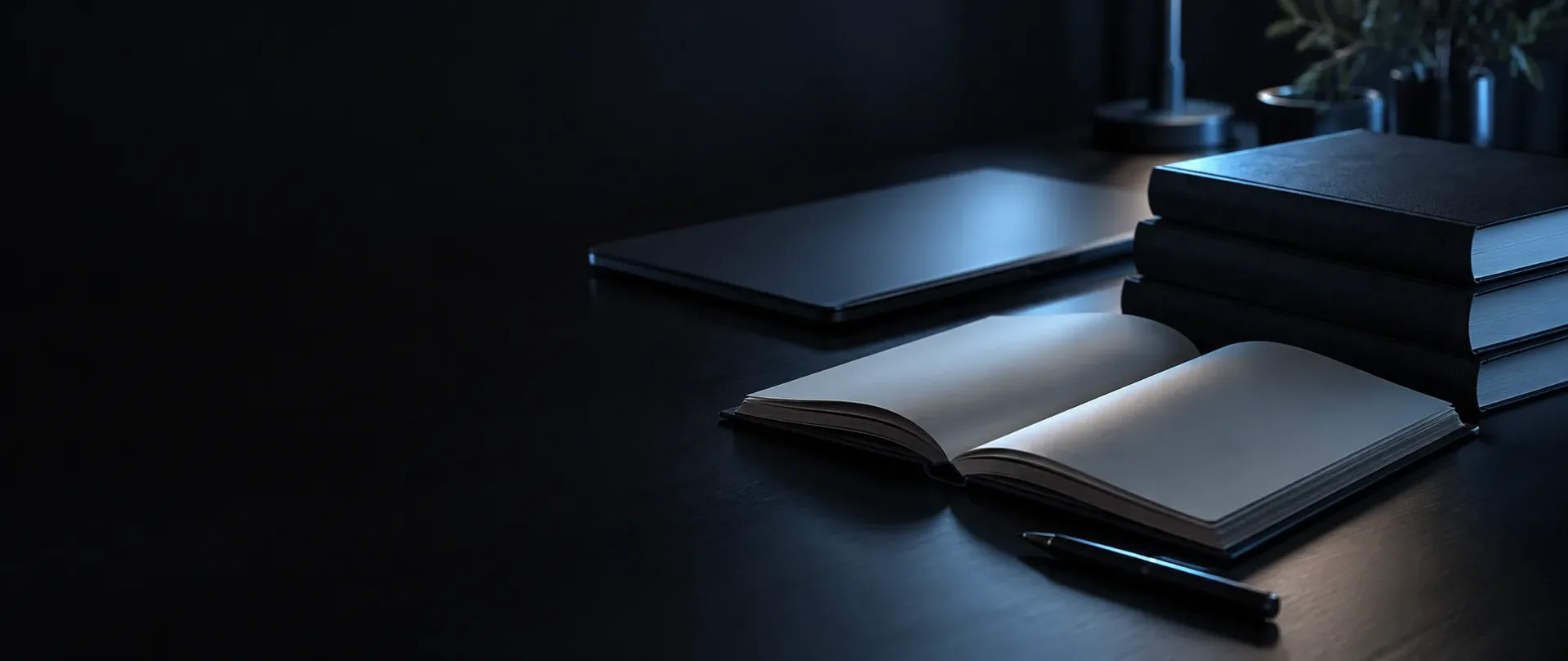 Dark desk with a stack of closed reference books, an open blank notebook, a pen and a closed laptop under soft blue light